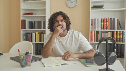 Man young indian writes in book with blue pen at desk with laptop, headphones and phone, bookshelves and clock behind in building  focused study. © Krakenimages.com