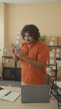 Man holding smartphone with hand covering mouth beside stacked cardboard boxes and laptop on desk in building office;excitement success.