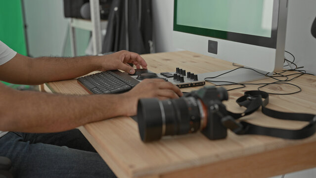 Man photographer seated at desk types on keyboard and turns editing dial with hands beside dslr camera and large monitor against green screen and shelves in studio; concentration.