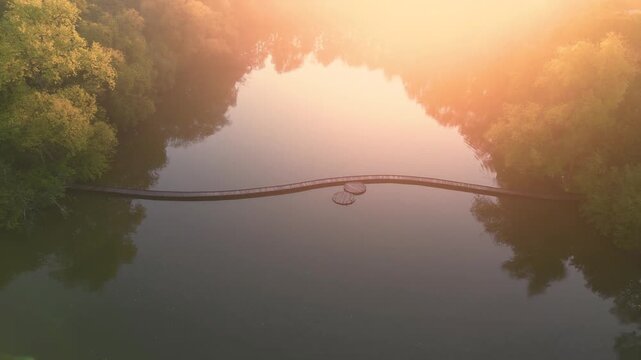 Sunset casts light on a lake with a wooden path. Trees surround the water. The wooden path leads across the lake, inviting exploration during dusk hour. Aerial drone footage