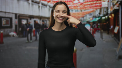 Fototapeta premium Woman in black long sleeve shirt points index finger to nose while smiling on a city street decorated with red lanterns and pedestrians; playful confidence.