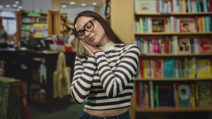 Woman wearing glasses and striped shirt rests head on hands pressed to cheek for sleep in a building library among bookshelves and reading tables  quiet relaxed study break. © Krakenimages.com