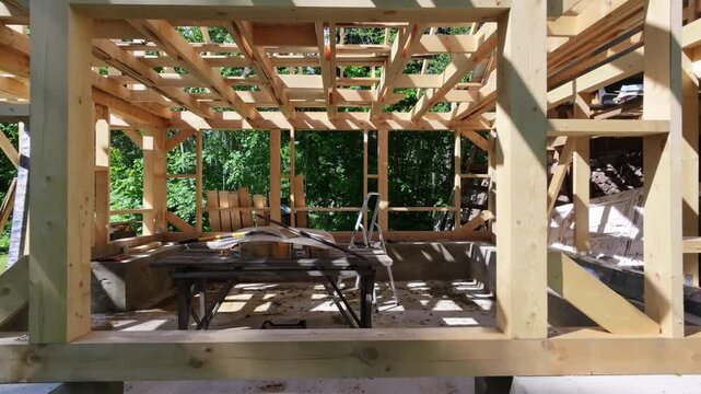 A timber frame stands on a stone base as work continues on a new structure. Vertical posts and crossbeams form the outline while tools, loose boards and a workbench rest inside the open space. 