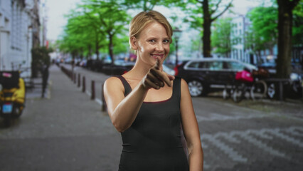 Fototapeta premium Woman pointing index finger on leafy city street wearing black tank top, smiling direct gesture toward camera; friendly invitation.
