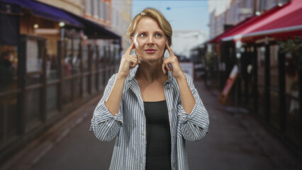 Fototapeta premium Woman in a striped shirt gesturing with open hands toward camera on a bustling street with sidewalk cafes, awnings and outdoor tables; thinking.