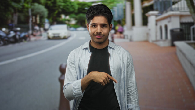 Young hispanic man points finger down at brick sidewalk on a street beside a bollard, wearing unbuttoned shirt and black tshirt; playful confidence.