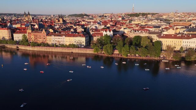 Aerial drone view of the Vltava river lined with small pedal boats, Prague old town waterfront with Tyn Church spires and red rooftops glowing in golden hour light, Prague, Czech Republic.