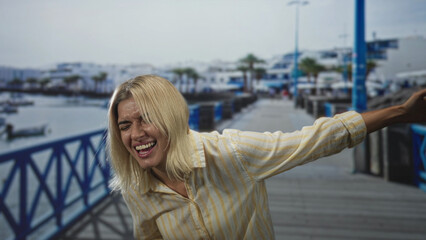 Obraz premium Woman pointing to her stomach and laughing with eyes closed, arm outstretched on railing on street by marina pier; joyful laughter.