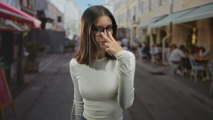 Obraz premium Woman wearing glasses points finger toward camera while standing near outdoor cafe tables on a street; confidence challenge.