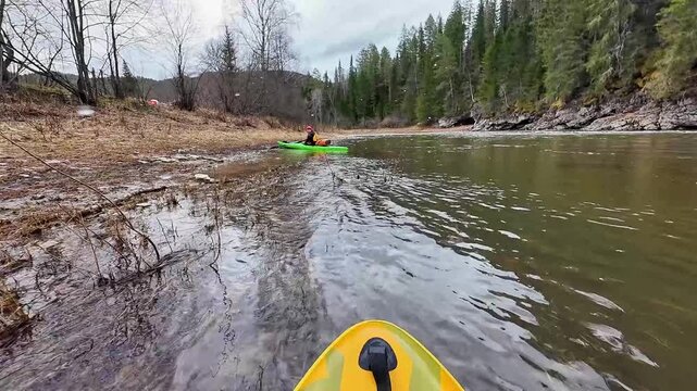 Kayak launch shallow river helper pushes green kayak from muddy bank, companion in yellow kayak films POV, jacket and red cap visible, rocky shore and pine forest, overcast sky, gentle