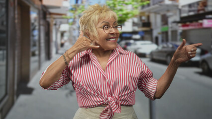 Senior woman making phone gesture with hand and pointing finger in a lively urban street scene  playful greeting joy. © Krakenimages.com