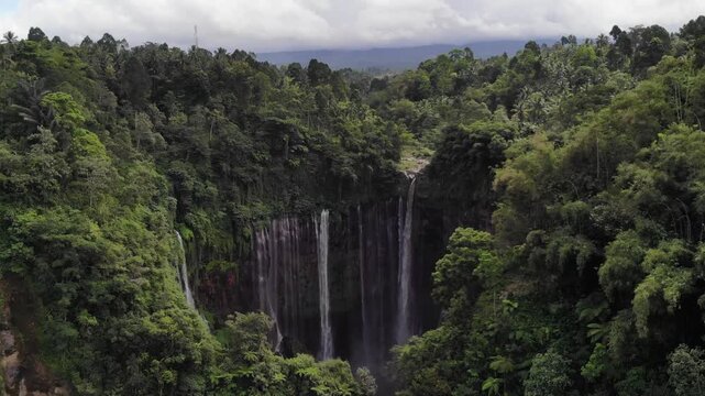 Low Aerial Drone Approaching Tumpak Sewu Waterfall Through Tropical Forest with Mountain Backdrop, Java, Indonesia