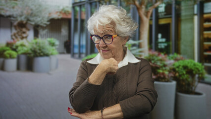 Fototapeta premium Elderly woman wearing colorful glasses points index finger and rests chin with manicured hand near large planters in front of building entrance; quiet confidence.