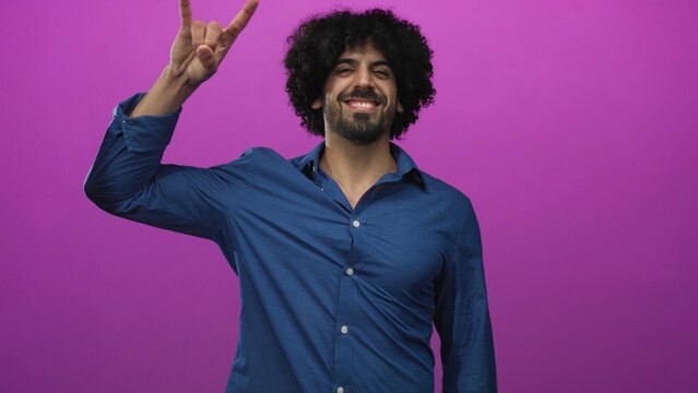 Curly haired man making rock horns gesture with raised hand and smiling in magenta studio; playful fun energy.