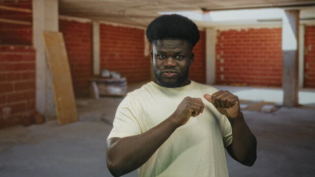 Young african american man points finger to chest and makes fist while looking assertive in unfinished building interior wearing white t shirt and beard; confidence.