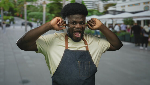 Man plugging fingers into ears on a busy street wearing denim apron and pale yellow t shirt; annoyance.