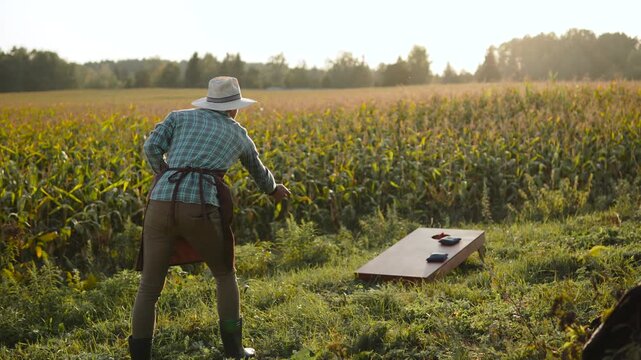 Farmer successfully playing in cornhole during break in corn field. Historical chronicle of cornhole game development. Agricultural worker successfully tossing corn bag into hole of wooden board.