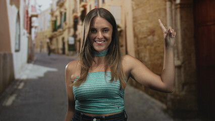 Naklejka premium Woman raises bare arm and points finger upward on a narrow street wearing turquoise crop top and choker; effortless confidence.