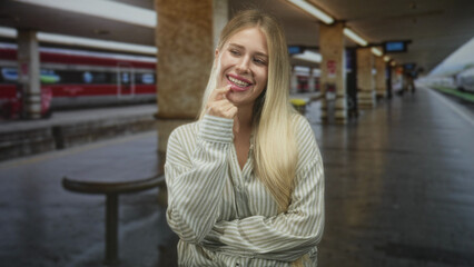 Fototapeta premium Woman blonde young touching lip with finger while smiling on train platform in building near marble pillars; cheerful wanderlust.