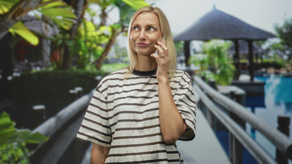 Fototapeta premium Woman holding smartphone to ear on resort pool walkway near gazebo building during phone call; quiet contemplation.