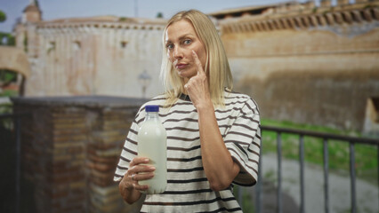 Fototapeta premium Woman holding milk bottle with clasped hands and steady gaze in front of an old stone building and iron railing; serenity domesticity.