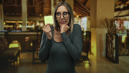 Fototapeta premium Woman with glasses and blonde hair holding a sad yellow postit note, hand under chin and biting lip in a hotel building lobby; anxiety.