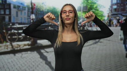 Fototapeta premium Young hispanic blonde woman pointing thumbs at chest on city street wearing red glasses and black top; self confidence.