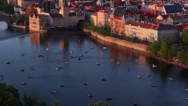 Aerial drone view of the Vltava river with pedal boats, Prague old town embankment featuring a clock tower and historic facades glowing in warm sunset light, Prague, Czech Republic.