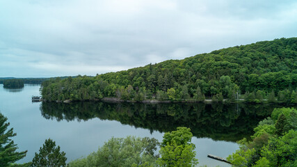Obraz premium A stunning high-angle drone photograph capturing the lush green forested hills of Muskoka perfectly reflecting like a mirror on the glassy, calm water of Lake of Bays under a cloudy spring sky.