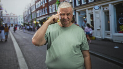 Obraz premium Man points finger to temple on a busy street near storefronts and sidewalk, wearing a green tshirt and standing center frame; thoughtful reflection.
