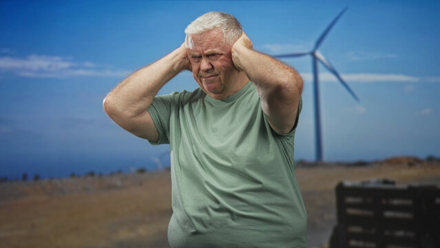 Senior man covering ears at a wind farm beside a large wind turbine and stacked crates on a dry coastal plain; noise annoyance.