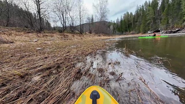 Kayak entering shallow reed filled marsh, paddler easing into grassy bank to land, calm slow water, muddy bottom and wetland vegetation, companion kayak visible on distant shore