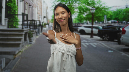Fototapeta premium Thai woman with hands on chest and fingers intertwined, wearing white sundress, standing on a city street with parked cars; gratitude.