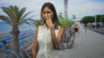 Fototapeta premium Woman pointing index finger to camera on beach promenade wearing sleeveless white shirt, smiling and gesturing with right hand; playful confidence.