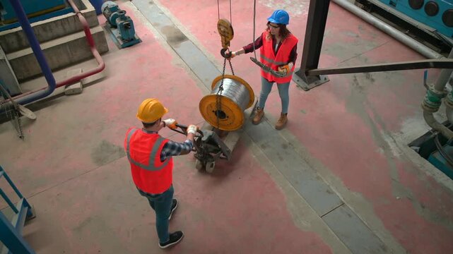 Two workers in hard hats and orange vests maneuver a large spool of cable on a dolly in an industrial factory setting for installation or maintenance purposes	