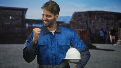 Fototapeta premium Man construction worker wearing blue overalls, holding white hardhat with beard visible, smiling and fist pump in front of stone ruins building; pride duty.