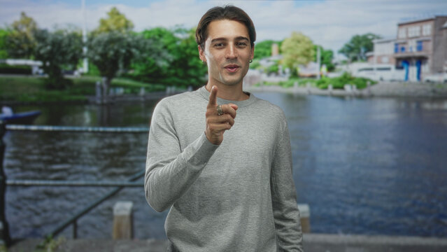 Man pointing finger and gesturing with hands over chest beside a riverside building at a canal quay while addressing the camera; friendly gratitude.