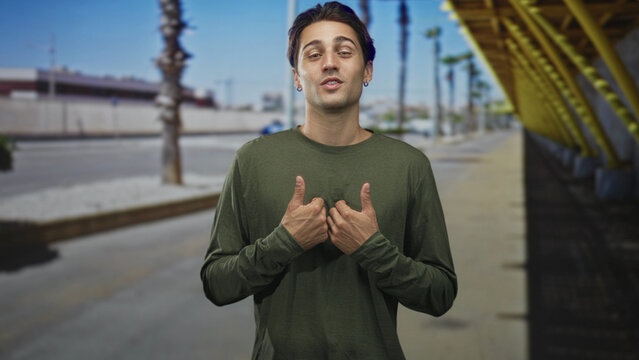 Young man with thumbs to chest, smirking and speaking at a street bus stop under a yellow canopy while wearing a green longsleeve shirt; sheepish doubt.