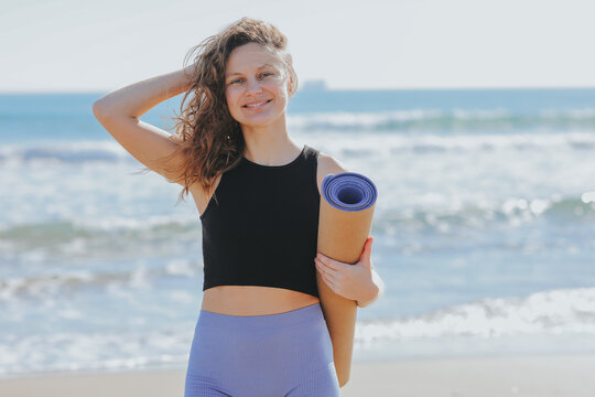 Young woman holds yoga mat, looks at camera and smiles. Lady prepares to workout outdoors. Sport and fitness on seashore. Healthy lifestyle, morning exercises, female wellness and power, healthcare.