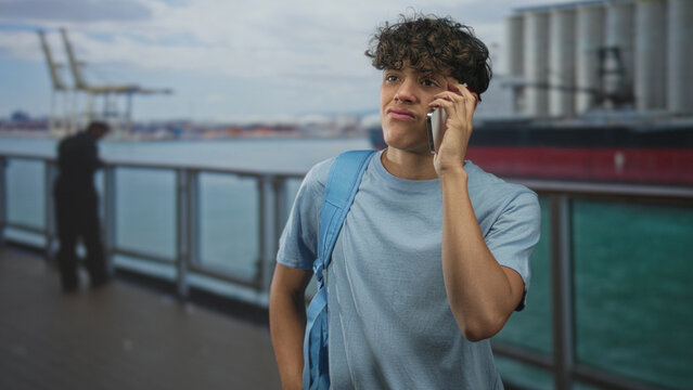 Teenage boy with backpack holding phone to ear on street pier beside cargo ship, eyes half closed and lips pursed midcall; impatience waiting.