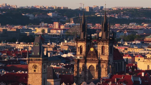 Aerial drone view of the Old Town Square with Tyn Church twin gothic towers and the Old Town Hall clock tower above red rooftops at sunset.