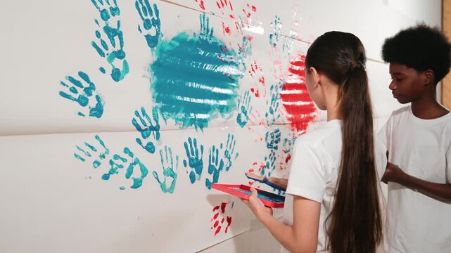 Side view of young children stamp hand with color and paint wall. high school student with mixed raced holding tray while paint room with red and blue in art lesson. Creative activity. Edification