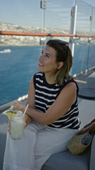 Obraz premium Woman sipping cocktail on cruise terrace, hand holding glass with pineapple garnish and sunglasses on head while seated on sofa; summer getaway relaxation.