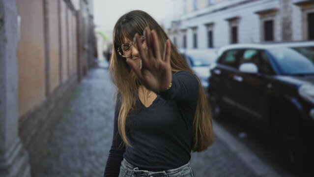 Woman with glasses raises hand to block camera, showing hand and necklace on a cobbled street beside a parked car; shyness privacy.
