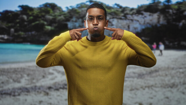 Young black man with glasses puffs cheeks and points to face on a sandy beach in a yellow sweater; playful expression.