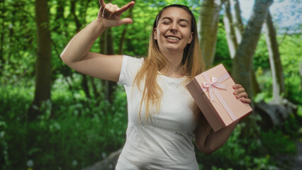 Fototapeta premium Woman holding pink present with bow while making a rock horns gesture and tilting head back in a forest; joyful celebration.