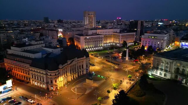 Cinematic aerial drone shot of Revolution Square in Bucharest, Romania at night. Beautiful city lights, heavy street traffic, and historic architectural landmarks under the night sky.