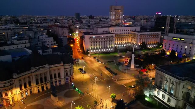 Cinematic aerial drone shot of Revolution Square in Bucharest, Romania at night. Beautiful city lights, heavy street traffic, and historic architectural landmarks under the night sky.