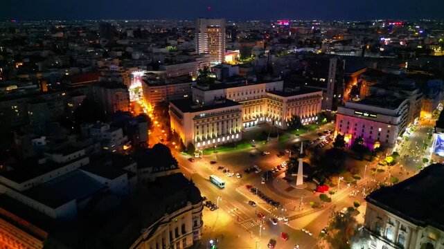 Cinematic aerial drone shot of Revolution Square in Bucharest, Romania at night. Beautiful city lights, heavy street traffic, and historic architectural landmarks under the night sky.