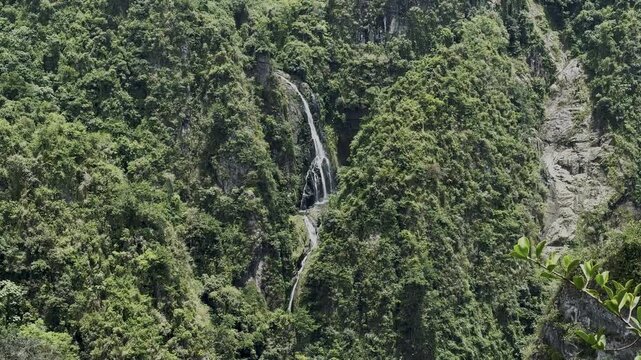 San Cristobal Canyon in Barranquitas, Puerto Rico, shows a tall waterfall flowing down steep rocky cliffs covered in dense tropical greenery. The canyon walls are rich with layered foliage, deep green